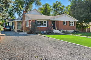 Ranch-style home with brick siding, a front lawn, gravel driveway, a chimney, and a shingled roof