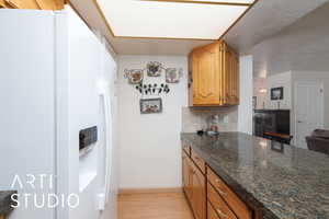 Kitchen with white refrigerator with ice dispenser, light wood-style floors, decorative backsplash, brown cabinets, and dark stone countertops