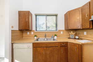 Kitchen with white dishwasher, light countertops, backsplash, and brown cabinetry