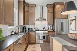 Kitchen with high quality appliances, wall chimney range hood, tasteful backsplash, dark wood-style flooring, and brown cabinetry