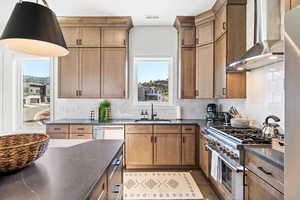 Kitchen featuring wall chimney exhaust hood, stainless steel stove, backsplash, brown cabinetry, and dark stone countertops