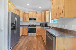 Kitchen featuring light brown cabinetry, appliances with stainless steel finishes, dark countertops, dark wood-type flooring, and under cabinet range hood