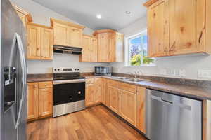 Kitchen featuring light brown cabinetry, appliances with stainless steel finishes, dark countertops, light wood-style flooring, and under cabinet range hood