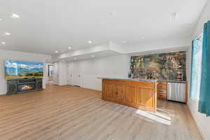 Indoor wet bar with brown cabinetry, light wood finished floors, stainless steel dishwasher, a glass covered fireplace, and recessed lighting