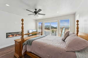 Bedroom featuring access to outside, dark wood-style flooring, a textured ceiling, a glass covered fireplace, and a ceiling fan