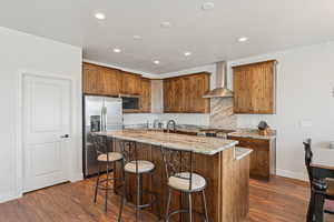 Kitchen with light stone counters, dark wood-type flooring, stainless steel appliances, wall chimney range hood, and a center island with sink