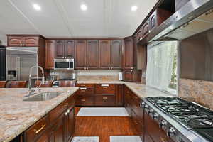 Kitchen with appliances with stainless steel finishes, dark brown cabinets, wall chimney exhaust hood, dark wood-style flooring, and light stone counters