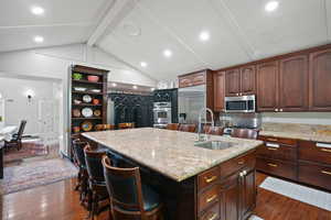 Kitchen with a breakfast bar area, dark wood-style flooring, dark brown cabinetry, an island with sink, and light stone countertops