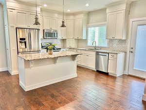 Kitchen with appliances with stainless steel finishes, light stone countertops, a kitchen island, dark wood-type flooring, and tasteful backsplash