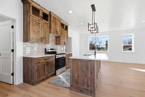 Kitchen with stainless steel electric range oven, decorative light fixtures, a kitchen island with sink, brown cabinets, and tasteful backsplash