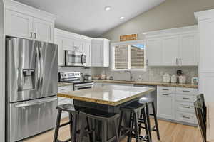 Kitchen with stainless steel appliances, light stone countertops, lofted ceiling, white cabinets, and a kitchen bar