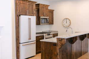 Kitchen with white appliances, light stone counters, light wood-style flooring, dark brown cabinets, and a breakfast bar