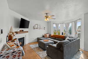 Living area featuring light wood finished floors, a glass covered fireplace, a textured ceiling, and a ceiling fan