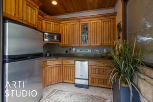 Kitchen with stainless steel appliances, brown cabinets, dark countertops, glass insert cabinets, and wood ceiling