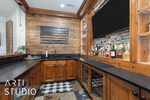 Indoor wet bar with brown cabinets, ornamental molding, open shelves, decorative light fixtures, and dark stone counters