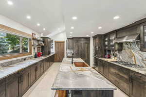 Kitchen featuring dark brown cabinets, light stone countertops, a center island with sink, vaulted ceiling, and recessed lighting