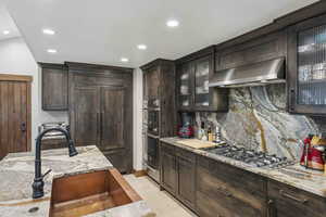 Kitchen featuring dark brown cabinetry, light stone counters, backsplash, ventilation hood, and recessed lighting