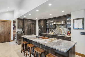 Kitchen with dark brown cabinets, glass insert cabinets, a kitchen breakfast bar, light stone countertops, and a center island with sink