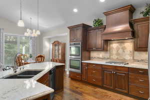Kitchen featuring premium range hood, light stone counters, dark wood-type flooring, hanging light fixtures, and appliances with stainless steel finishes
