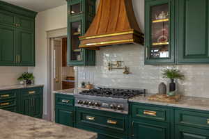 Kitchen featuring green cabinets, decorative backsplash, custom range hood, stainless steel gas cooktop, and glass insert cabinets