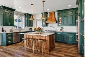 Kitchen with green cabinetry, light stone counters, tasteful backsplash, premium range hood, and appliances with stainless steel finishes