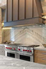 Kitchen with premium range hood, range with two ovens, light stone counters, and backsplash