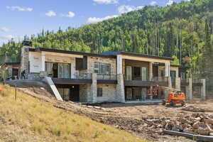 Rear view of house with stone siding, a balcony, a patio, a chimney, and a wooded view