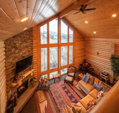 Living room with high vaulted ceiling, rustic walls, a stone fireplace, wood ceiling, and a ceiling fan