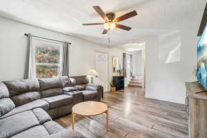 Living area with a textured ceiling, light wood-type flooring, ceiling fan, and stairway