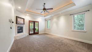 Carpeted empty room featuring french doors, crown molding, a raised ceiling, ceiling fan, and a fireplace