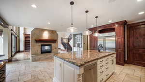 Kitchen featuring dark stone counters, an island with sink, stone tile floors, decorative light fixtures, and appliances with stainless steel finishes