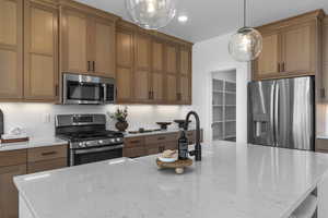 Kitchen with pendant lighting, stainless steel appliances, light stone counters, an island with sink, and backsplash