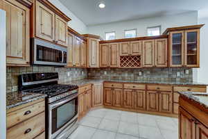 Kitchen with appliances with stainless steel finishes, backsplash, light tile patterned floors, dark stone counters, and glass insert cabinets