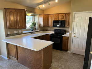 Kitchen with brown cabinetry, black appliances, a peninsula, light countertops, and vaulted ceiling