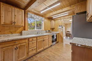 Kitchen with appliances with stainless steel finishes, light wood-style floors, wood walls, a wooden ceiling with exposed beams, and light stone countertops