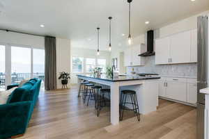 Kitchen featuring a kitchen island with sink, open floor plan, a kitchen bar, white cabinetry, and wall chimney exhaust hood