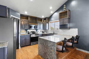 Kitchen with vaulted ceiling, open shelves, light wood finished floors, appliances with stainless steel finishes, and light stone counters