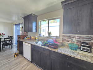 Kitchen with dark brown cabinets, white dishwasher, light wood-type flooring, and light stone countertops