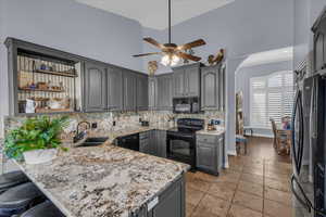 Kitchen with gray cabinetry, tasteful backsplash, black appliances, a peninsula, and light stone countertops