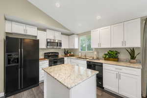 Kitchen with black appliances, white cabinetry, vaulted ceiling, light stone counters, and dark wood-style floors