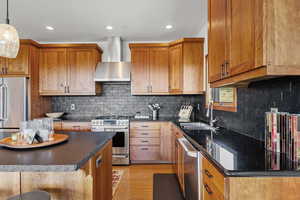 Kitchen featuring appliances with stainless steel finishes, wall chimney exhaust hood, brown cabinetry, light wood finished floors, and recessed lighting