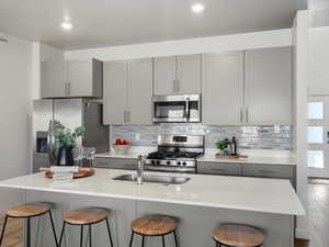 Kitchen with gray cabinetry, light wood-type flooring, appliances with stainless steel finishes, tasteful backsplash, and a center island with sink