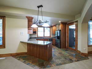 Kitchen with brown cabinetry, stone tile floors, black fridge, a peninsula, and dark stone counters