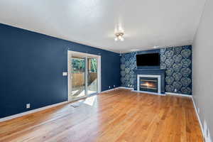 Unfurnished family room featuring light wood finished floors, fireplace, wallpapered  accent wall, and sliding glass door out outdoor covered patio.