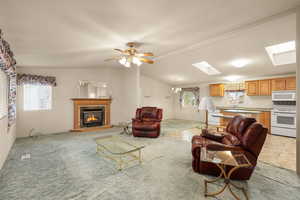 Living room with vaulted ceiling, a glass covered fireplace, a skylight, light carpet, and ceiling fan