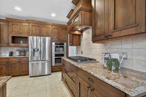 Kitchen featuring stainless steel appliances, brown cabinetry, light stone countertops, light tile patterned flooring, and recessed lighting