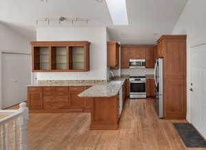 Kitchen featuring brown cabinetry, a skylight, appliances with stainless steel finishes, light wood finished floors, and light stone countertops