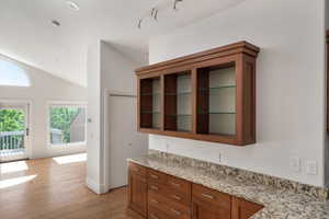Kitchen with lofted ceiling, brown cabinetry, light stone countertops, glass insert cabinets, and light wood-style floors