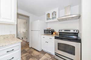 Kitchen with stainless steel appliances, white cabinetry, wall chimney range hood, light stone counters, and glass insert cabinets