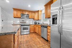 Kitchen featuring appliances with stainless steel finishes, dark stone countertops, light tile patterned flooring, recessed lighting, and brown cabinetry
